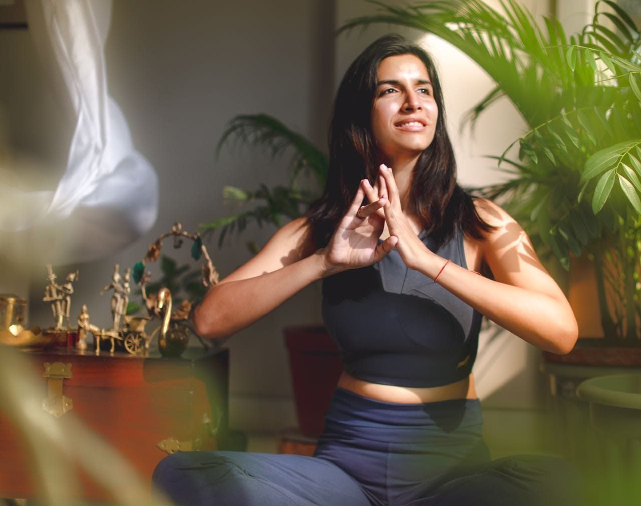 Varuna in seated meditation with hands in Anjali mudra, surrounded by plants and Indian brass figurines
