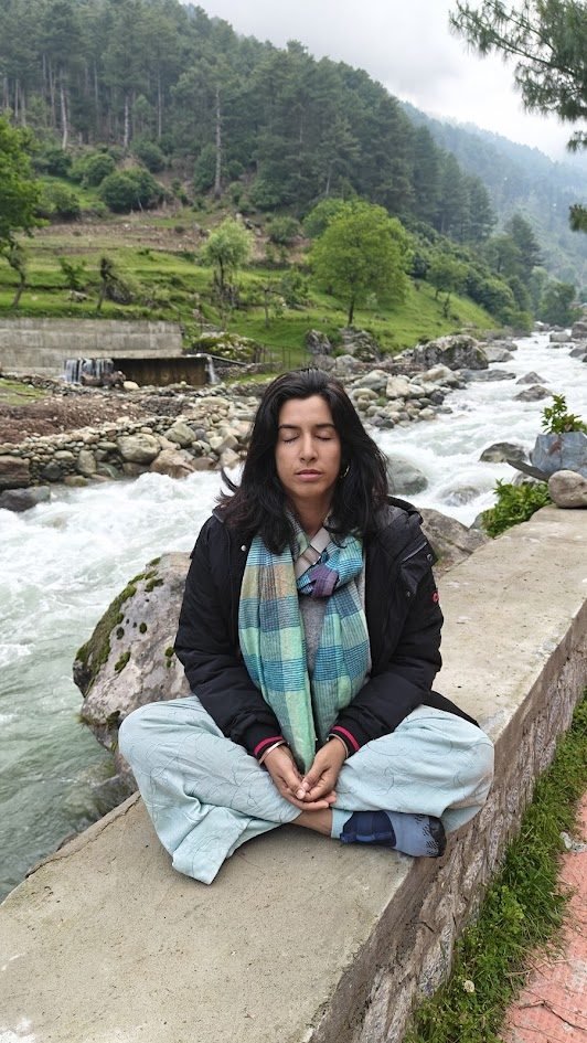 Varuna Shunglu meditating in lotus position on a stone wall by a fast-flowing Kashmiri river, surrounded by green forest hills