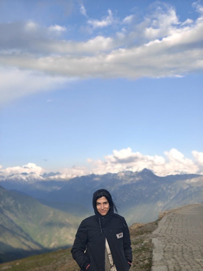 Varuna Shunglu standing on a high-altitude mountain road with dramatic Himalayan cloud sky behind her
